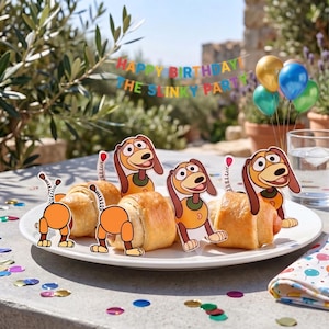 May include: A white plate holds several crescent roll appetizers decorated with paper cutouts of a cartoon dog character. The cutouts feature the dog's head, body, and spring-like tail. A banner in the background reads HAPPY BIRTHDAY THE SLINKY PARTY.