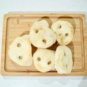 May include: A wooden cutting board holds several pale, round, baked goods with small holes. The baked goods are light in color with darker spots. The cutting board is made of light brown wood.