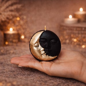 May include: A black and gold moon-shaped candle with a face design. The candle is round and has a gold crescent moon on one side and a black face on the other. The candle is held in a hand.