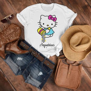 以下が含まれることがあります： 白いTシャツに漫画の猫のデザインと「Aquarius」の文字。画像には、デニムショーツ、茶色のカウボーイブーツ、麦わら帽子、茶色の革のバッグ、サングラスが含まれています。