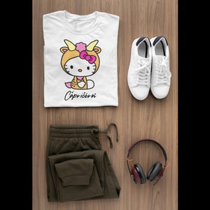 May include: A flat lay featuring a white t-shirt with a Capricorn graphic, olive green joggers, white trainers, and maroon headphones. The t-shirt displays a cartoon character in a Capricorn costume. The word "Capricorn" is written in cursive.