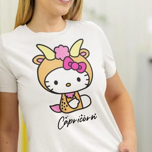 May include: White t-shirt with a cartoon character in a Capricorn costume. The character has a tan goat outfit with yellow horns, a pink bow, and the word "Capricorn" in black script.