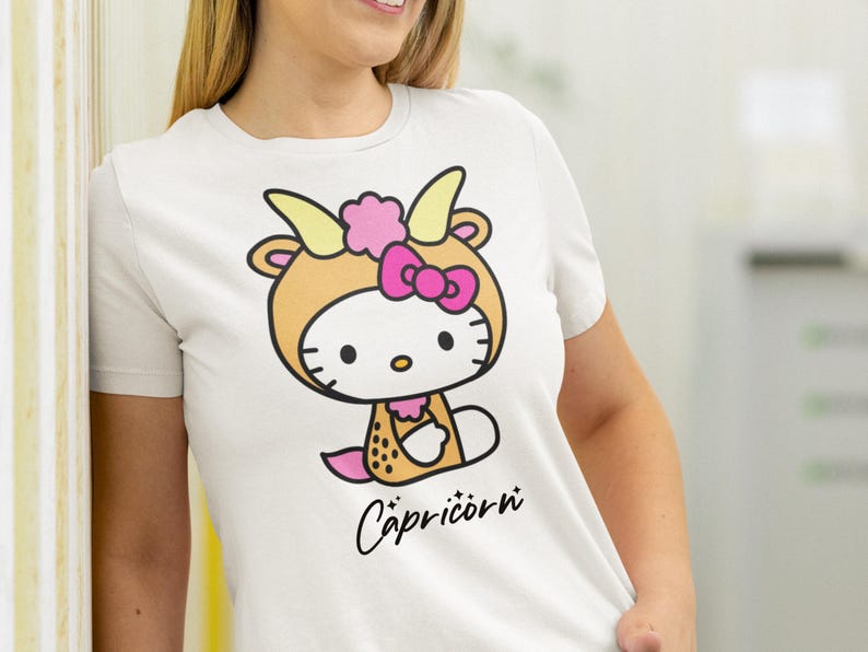 May include: White t-shirt with a cartoon character in a Capricorn costume. The character has a tan goat outfit with yellow horns, a pink bow, and the word "Capricorn" in black script.