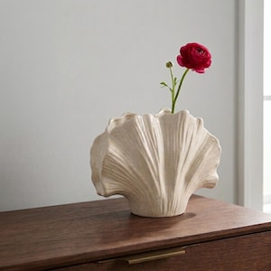 May include: A cream-colored, seashell-shaped ceramic vase with a single red flower. The vase has a textured surface and is displayed on a dark wooden surface. The flower has a green stem and leaves.