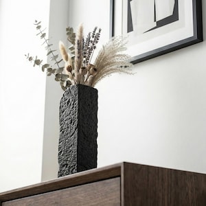 Puede incluir: Un jarrón rectangular negro texturizado lleno de flores y hierbas secas. El jarrón tiene una apariencia rugosa, similar a la piedra, y se coloca sobre una superficie de madera oscura. El arreglo incluye eucalipto, lavanda y hierba de la pampa.