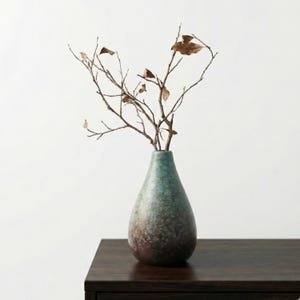 Può includere: Un vaso a forma di goccia con finitura screziata verde acqua e marrone, contenente diversi rami secchi con piccole foglie marroni. Il vaso è posto su una superficie di legno scuro su sfondo bianco.