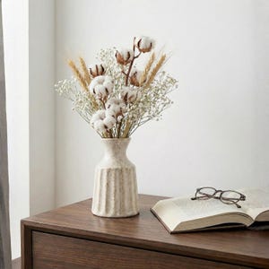 Könnte beinhalten: Eine beige Keramikvase mit vertikalen Rillen enthält einen Strauß aus Baumwollblüten, Weizenähren und weißem Schleierkraut. Die Vase steht auf einer dunklen Holzoberfläche neben einem offenen Buch und einer Brille. Das Arrangement hat eine natürliche, rustikale Ästhetik.