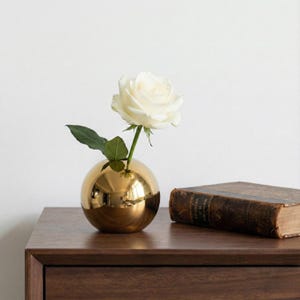 Op de afbeelding: Een gouden, bolvormige vaas met een enkele witte roos en groene bladeren. De vaas staat op een donkerhouten oppervlak naast een vintage boek met een bruine kaft.