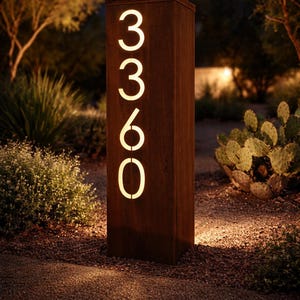 May include: A rectangular, illuminated address sign with the numbers 3360. The sign is made of a dark, weathered metal and is set in a garden with trees and shrubs. The numbers glow with a warm, inviting light.