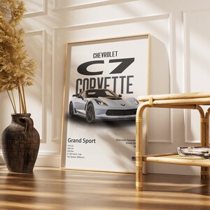May include: A framed poster featuring a silver Chevrolet Corvette Grand Sport. The poster has the car's name in large black letters and includes specifications. The poster is displayed in a room with a decorative vase and a wooden side table.