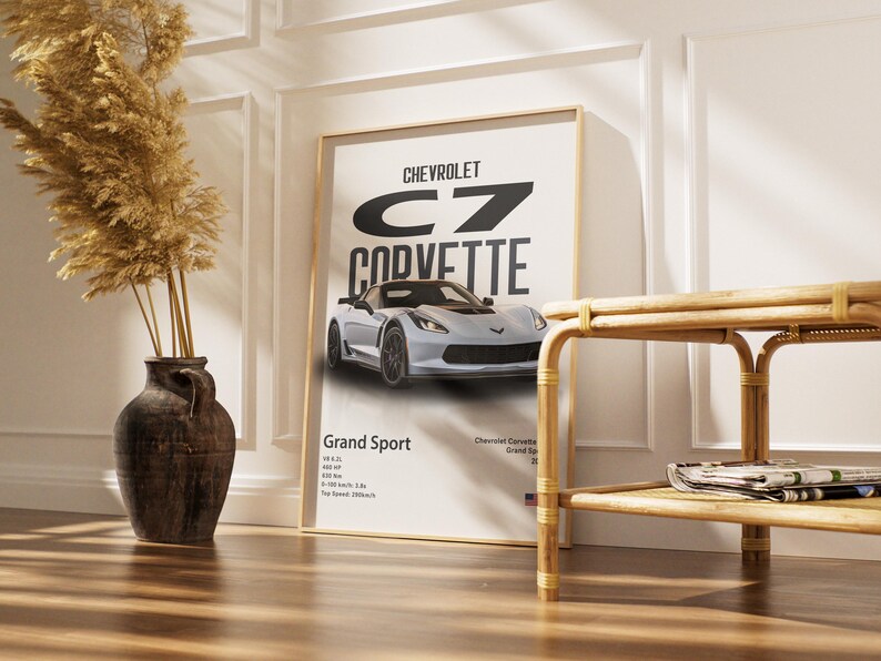 May include: A framed poster featuring a silver Chevrolet Corvette Grand Sport. The poster has the car's name in large black letters and includes specifications. The poster is displayed in a room with a decorative vase and a wooden side table.