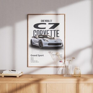 May include: A framed poster featuring a silver Chevrolet C7 Corvette Grand Sport. The poster includes the car's name, model, and specifications. The poster is displayed on a white wall above a wooden surface.