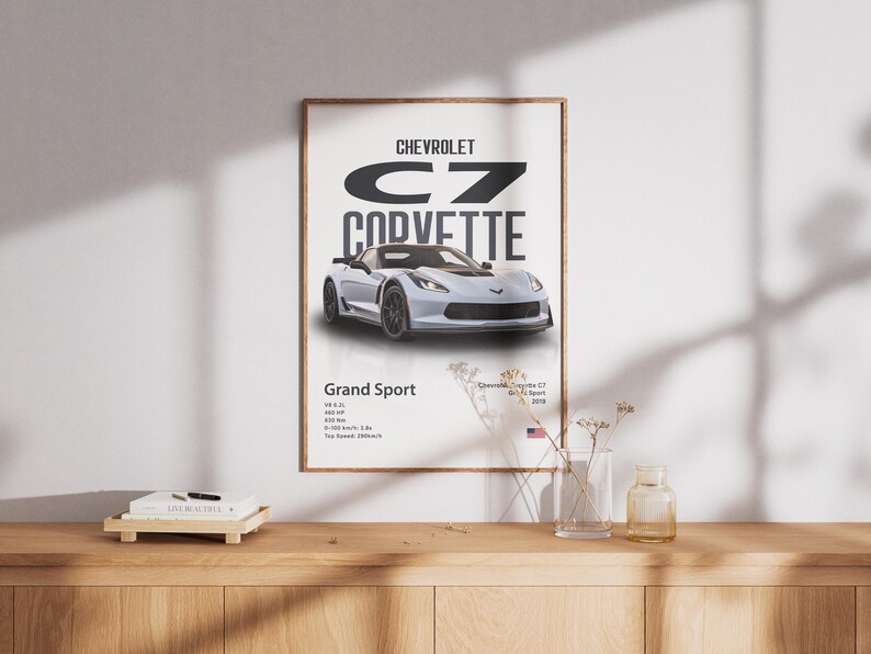 May include: A framed poster featuring a silver Chevrolet C7 Corvette Grand Sport. The poster includes the car's name, model, and specifications. The poster is displayed on a white wall above a wooden surface.