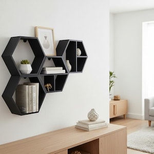 May include: Black hexagonal wall shelves displaying books, a small plant, and a framed picture. The shelves are mounted on a white wall, offering a modern and stylish storage solution for a living space.