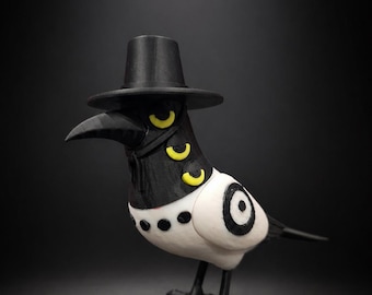 May include: A stylized bird figurine wearing a black top hat. The bird has a black head and beak, a white body, and black legs. It features three yellow crescent-shaped markings on its neck, a row of black dots on its chest, and a black target symbol.