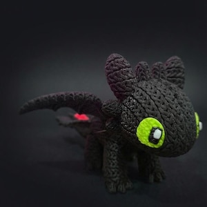 May include: A black dragon figurine with bright green eyes and small white highlights. The dragon has detailed wings and a textured body. The wings have a red accent. The dragon is a fantasy-themed item.