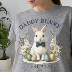 Peut inclure: T-shirt gris avec un motif de lapin blanc portant un nœud papillon bleu, entouré de fleurs et d'œufs de Pâques. Le texte "DADDY BUNNY" est arqué au-dessus du lapin, avec "EST. 2026" en dessous.