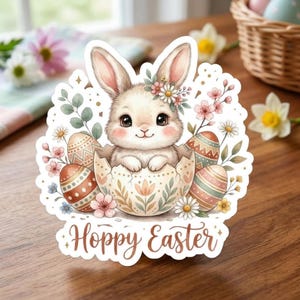 Peut inclure: Un autocollant représentant un mignon lapin sortant d'un œuf fêlé, entouré d'œufs de Pâques décorés et de fleurs. Le lapin porte une couronne florale. Les mots "Hoppy Easter" sont écrits en bas dans une police décorative.