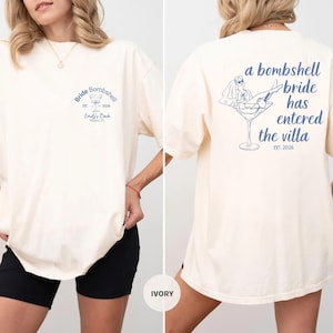 May include: An ivory t-shirt featuring blue graphics. The front displays a martini glass icon with the text Bride Bombshell Est. 2026 Emily's Bach Miami, FL. The back shows a bride in a veil lounging in a martini glass with the text a bombshell bride has entered the villa EST. 2026.