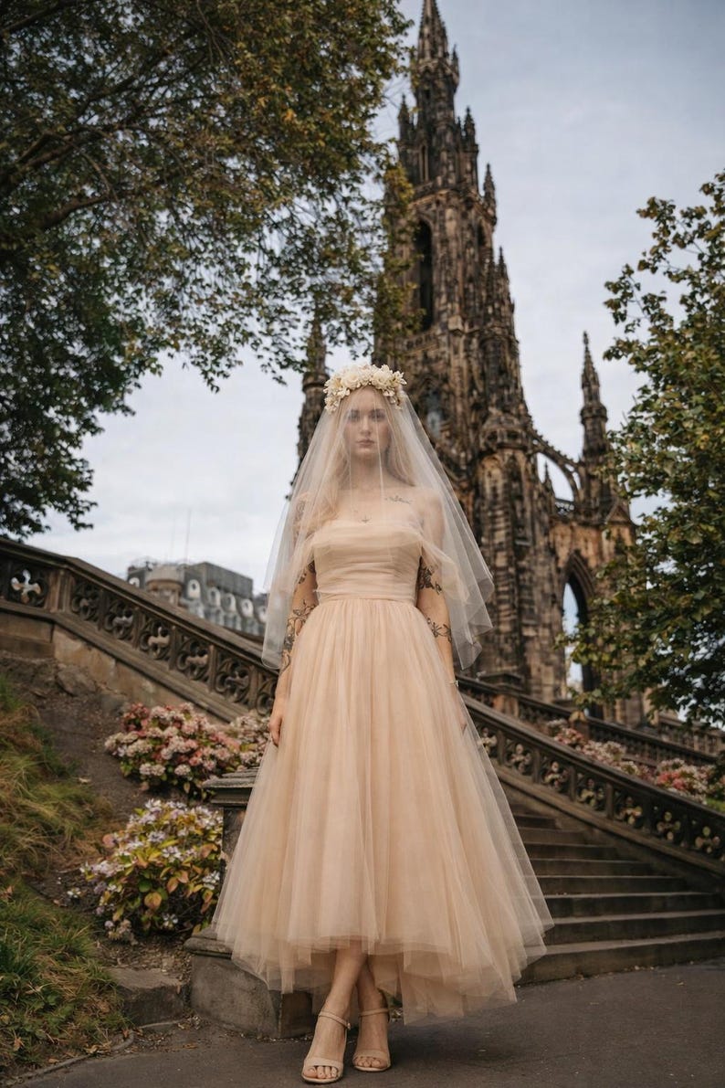 May include: A woman in a blush-colored tulle wedding dress with a floral crown and veil stands on stone steps. The dress is tea-length, and she wears strappy heels. A large stone building is in the background.