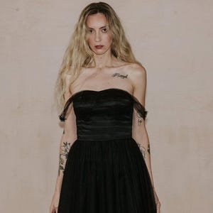 May include: A strapless black evening gown with a sweetheart neckline and a full tulle skirt. The dress features a fitted bodice and a flowing skirt. The model has long blonde hair and tattoos on their arms.