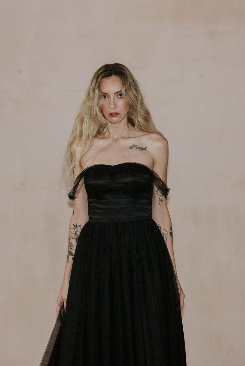 May include: A strapless black evening gown with a sweetheart neckline and a full tulle skirt. The dress features a fitted bodice and a flowing skirt. The model has long blonde hair and tattoos on their arms.