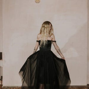 May include: A black off-the-shoulder dress with a tulle skirt. The dress has a fitted bodice and a flowing skirt. The model is wearing black platform heels. The background is a neutral wall with a light fixture.