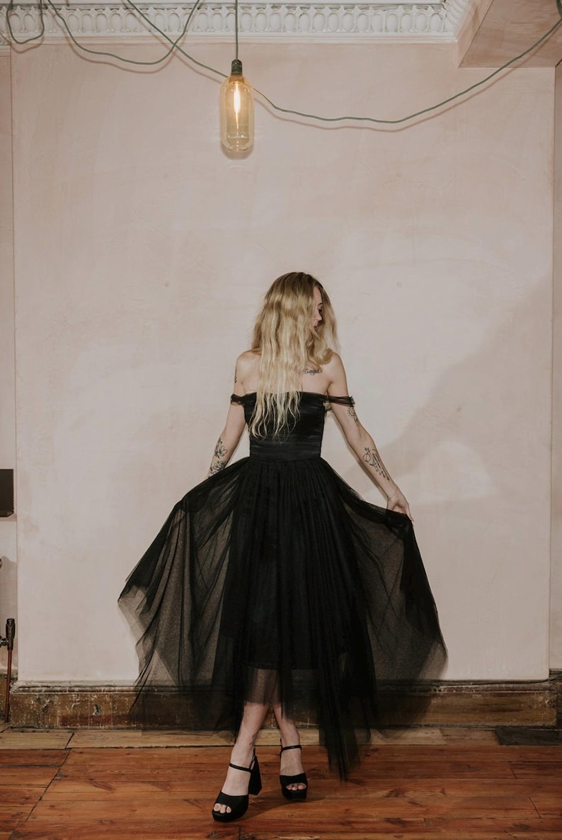 May include: A black off-the-shoulder dress with a tulle skirt. The dress has a fitted bodice and a flowing skirt. The model is wearing black platform heels. The background is a neutral wall with a light fixture.