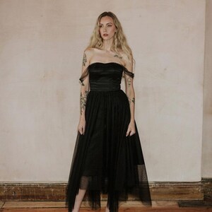 May include: A strapless black dress with a tulle skirt. The dress features a fitted bodice and a flowing skirt that falls to the mid-calf. The model is wearing black platform heels. The background is a neutral wall and wooden floor.