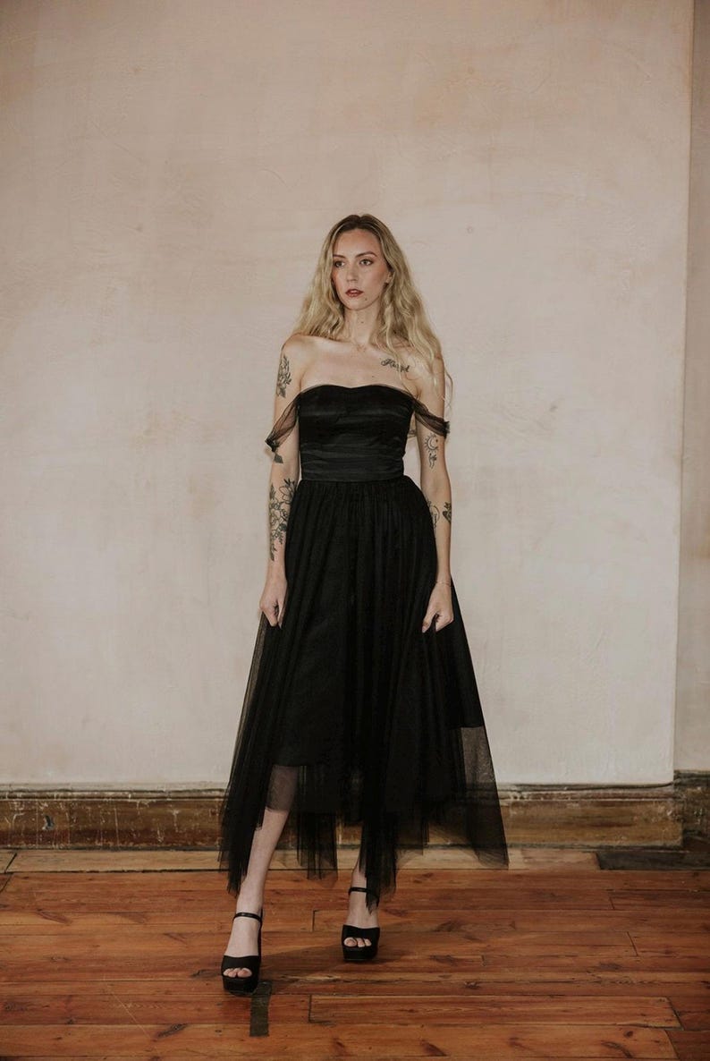 May include: A strapless black dress with a tulle skirt. The dress features a fitted bodice and a flowing skirt that falls to the mid-calf. The model is wearing black platform heels. The background is a neutral wall and wooden floor.