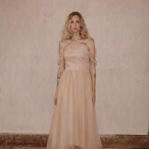 May include: A blush-colored, off-the-shoulder tulle dress. The dress features a fitted bodice and a flowing, high-low skirt. The model is wearing matching sandals. The background is a neutral, textured wall and a wooden floor.