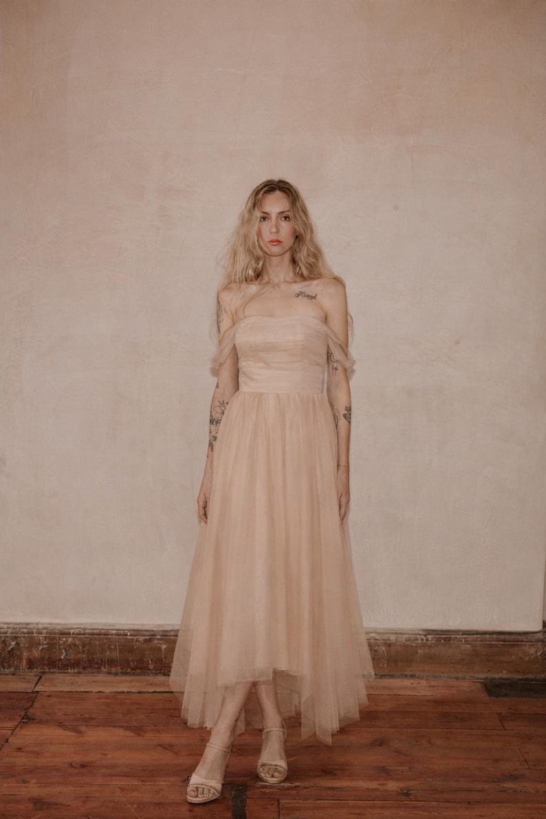 May include: A blush-colored, off-the-shoulder tulle dress. The dress features a fitted bodice and a flowing, high-low skirt. The model is wearing matching sandals. The background is a neutral, textured wall and a wooden floor.