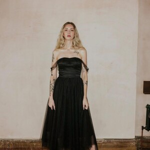 May include: A black strapless dress with a tulle skirt. The dress features a sweetheart neckline and a fitted bodice. The skirt is layered and falls to the mid-calf. The model is wearing black platform heels. The background is a neutral color.