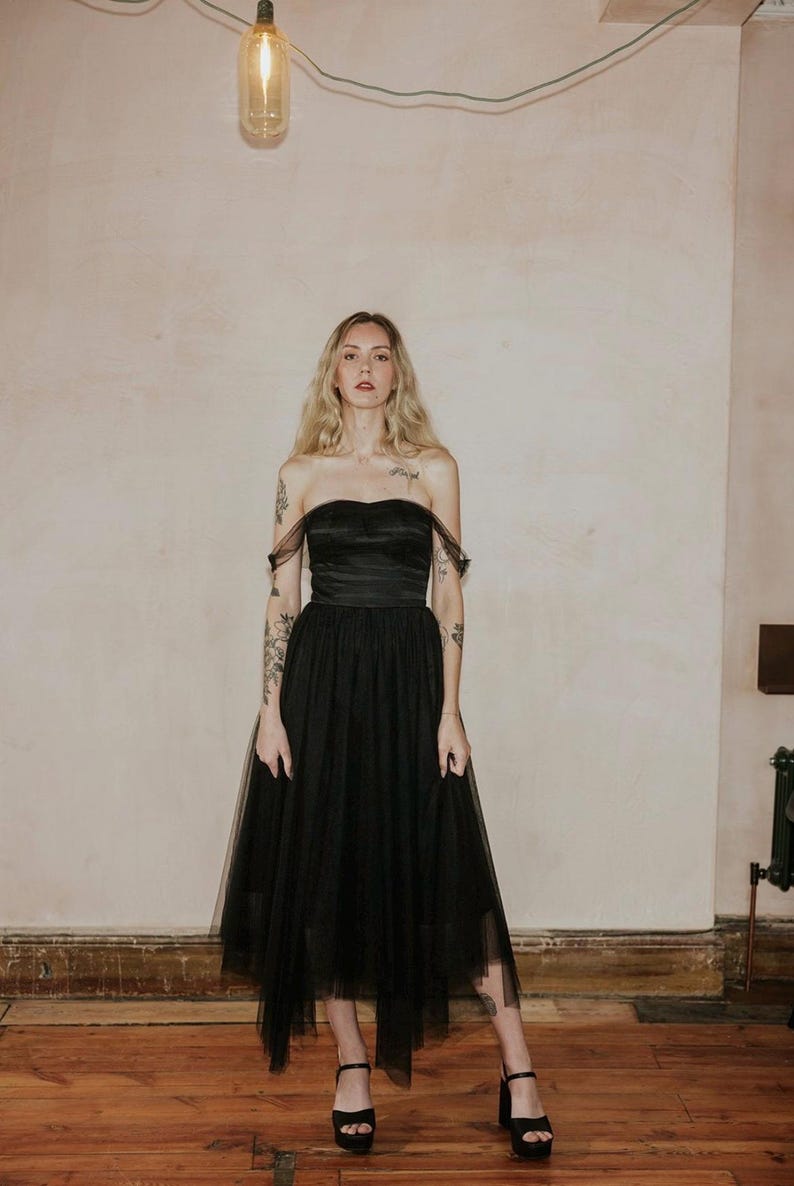 May include: A black strapless dress with a tulle skirt. The dress features a sweetheart neckline and a fitted bodice. The skirt is layered and falls to the mid-calf. The model is wearing black platform heels. The background is a neutral color.