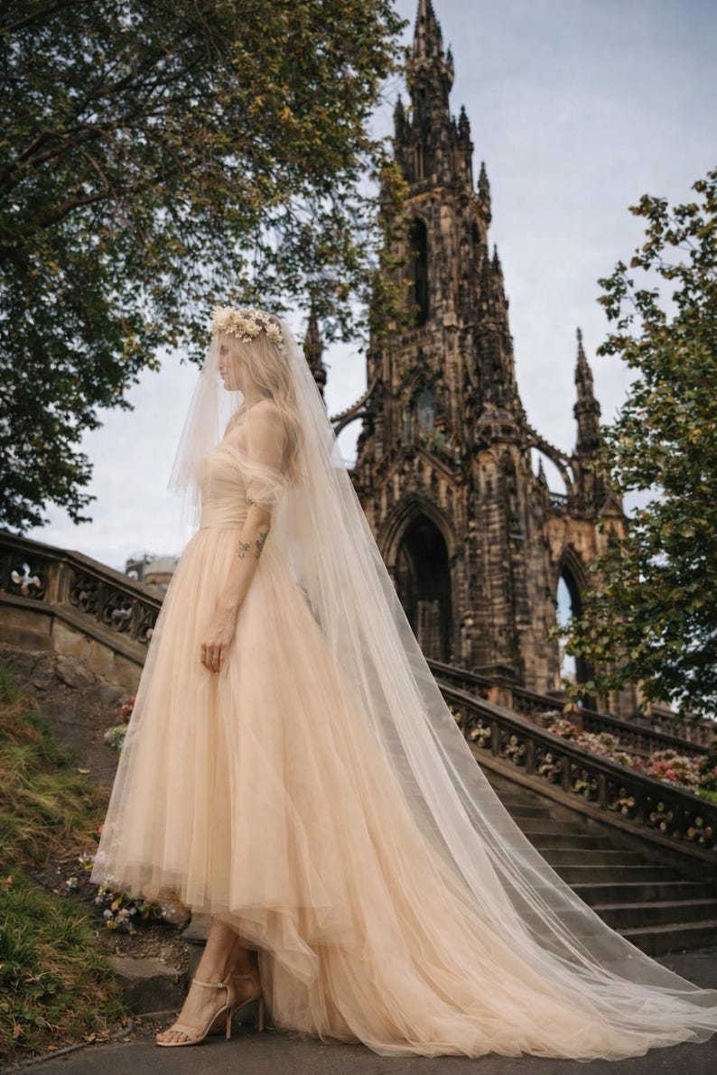 May include: A light peach-colored wedding dress with a tulle skirt and a long veil. The bride is wearing a floral crown and standing on a stone staircase in front of a large, ornate building. The dress has a high-low hemline.
