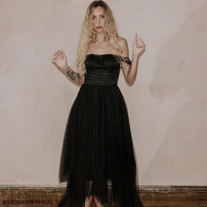 May include: A strapless black dress with a layered tulle skirt. The dress features a fitted bodice and a flowing, asymmetrical hemline. The model is wearing black platform heels.