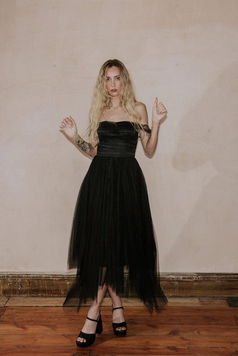 May include: A strapless black dress with a layered tulle skirt. The dress features a fitted bodice and a flowing, asymmetrical hemline. The model is wearing black platform heels.