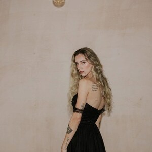 May include: A strapless black dress with a tulle skirt. The dress is worn by a person with long blonde hair and tattoos on their arms. A light bulb hangs from a wire in the background.
