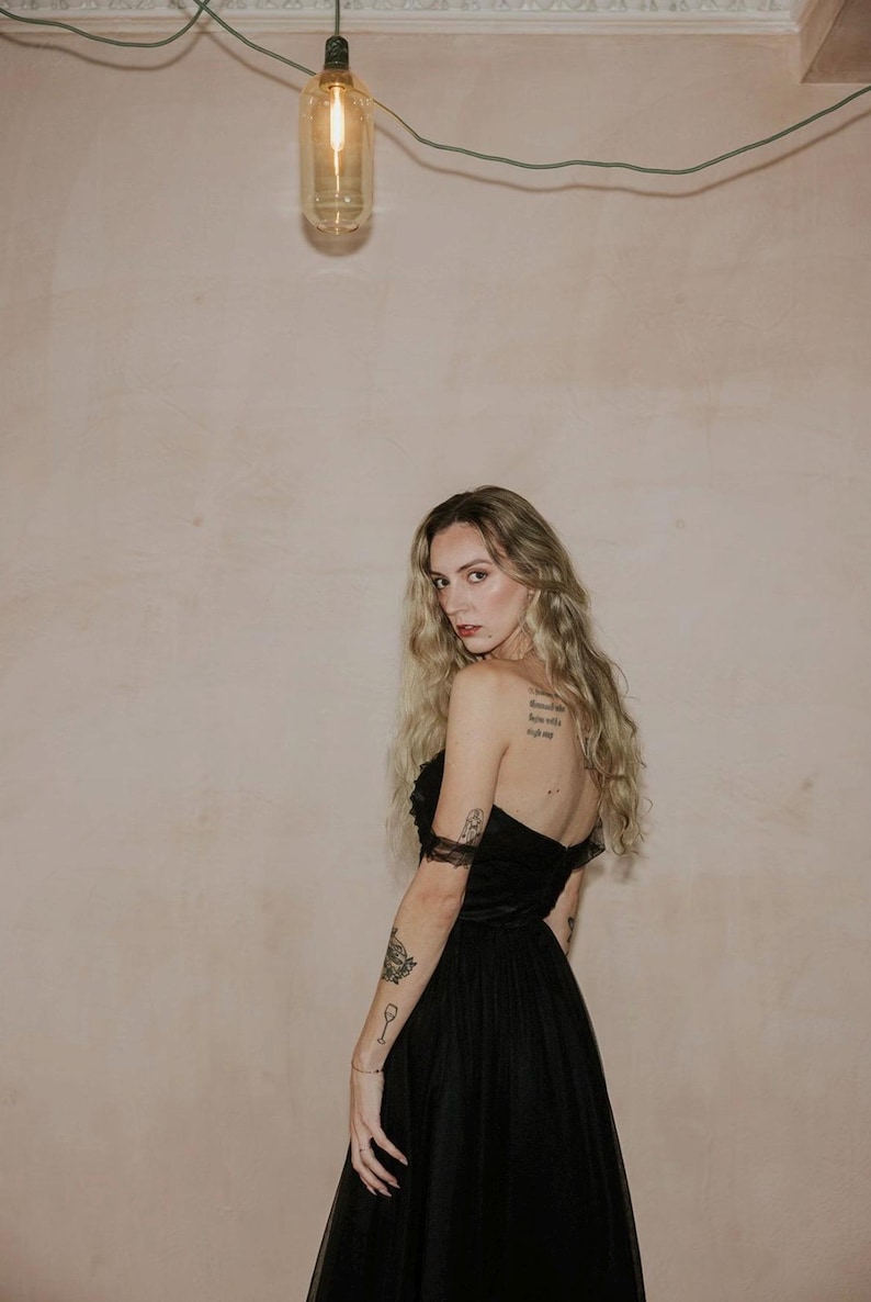 May include: A strapless black dress with a tulle skirt. The dress is worn by a person with long blonde hair and tattoos on their arms. A light bulb hangs from a wire in the background.