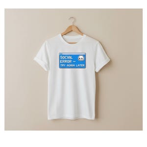 Op de afbeelding: Wit t-shirt met een blauwe afbeelding met de tekst "SOCIAL ERROR - TRY AGAIN LATER" en een gepixeleerd verdrietig gezicht. Het t-shirt hangt aan een houten hanger.