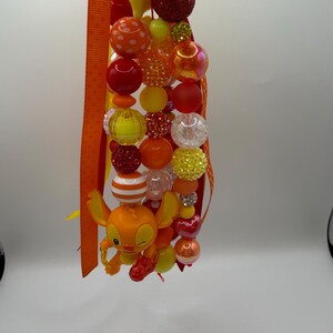 May include: A beaded lanyard featuring a variety of colorful beads in shades of orange, red, yellow, and clear. The lanyard includes a small orange character figure and is accented with orange ribbon.