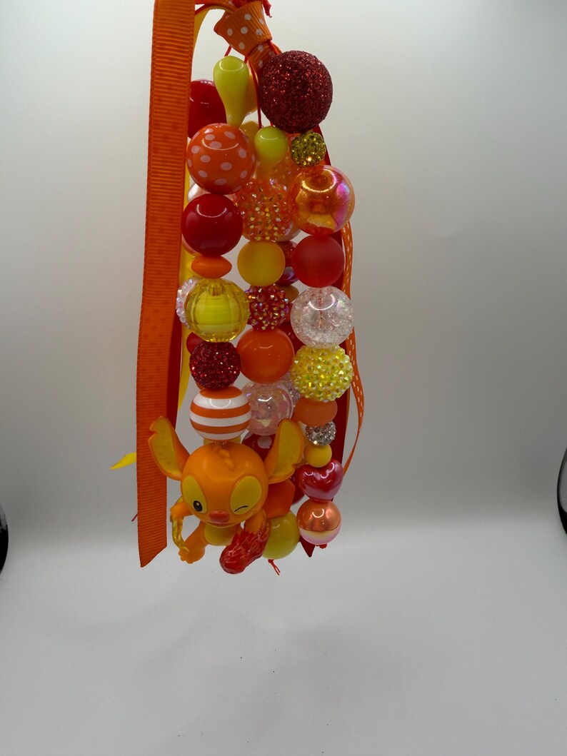 May include: A beaded lanyard featuring a variety of colorful beads in shades of orange, red, yellow, and clear. The lanyard includes a small orange character figure and is accented with orange ribbon.
