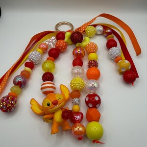 May include: A beaded keychain featuring a cartoon character in shades of orange and yellow. The keychain includes a variety of beads in red, orange, yellow, and white, with a gold-colored ring and orange ribbons.