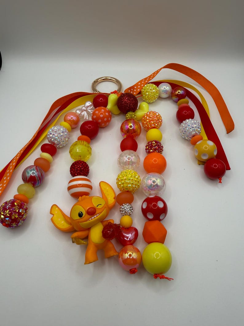 May include: A beaded keychain featuring a cartoon character in shades of orange and yellow. The keychain includes a variety of beads in red, orange, yellow, and white, with a gold-colored ring and orange ribbons.