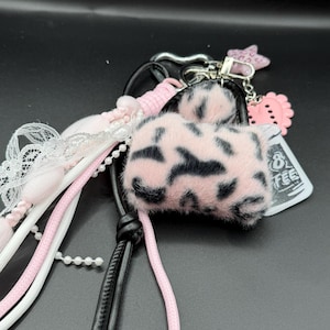 May include: A keychain featuring a miniature faux fur handbag with a pink and black leopard print, attached to a black cord, white lace, pink beads, and charms including a pink star, a pink heart, and a tag with the text 8 FEE.