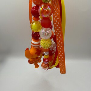May include: A beaded lanyard featuring a variety of colorful beads in shades of red, orange, yellow, and clear. The beads are interspersed with ribbons in orange, yellow, and red. A small orange figurine is attached to the lanyard.