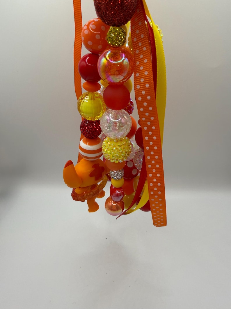 May include: A beaded lanyard featuring a variety of colorful beads in shades of red, orange, yellow, and clear. The beads are interspersed with ribbons in orange, yellow, and red. A small orange figurine is attached to the lanyard.