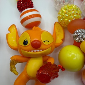 May include: An orange figurine of a cartoon character winking, holding a red object and a yellow object. The figurine is surrounded by colorful beads in red, white, orange, yellow, and clear.