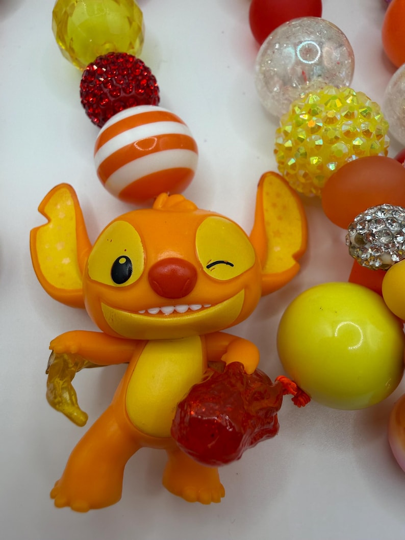 May include: An orange figurine of a cartoon character winking, holding a red object and a yellow object. The figurine is surrounded by colorful beads in red, white, orange, yellow, and clear.