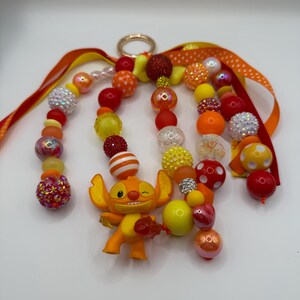 May include: A handmade keychain featuring an orange Stitch figurine and a variety of colorful beads in shades of red, orange, and yellow. The keychain includes a gold-colored ring and orange ribbons with polka dots.